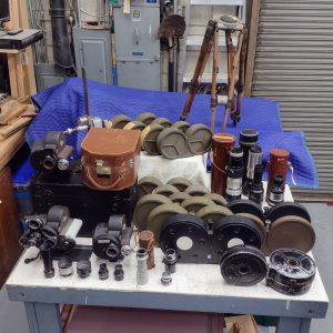 A picture of the recently acquired equipment lot, including3 Bell & Howell Eyemo cameras, some Bell & Howell magazines, some theodolite magazines, some Parvo magazines, a Mitchell tripod, and assorted lenses.