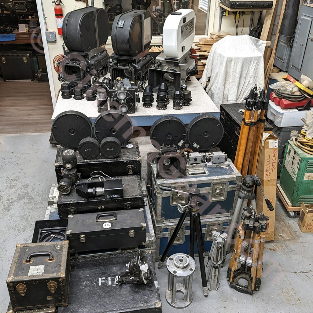 A full view of the collection of new camera equipment, including the rackover Mitchell BNC, the spinning mirror reflex Mitchell BNCR, the Mitchell 205, the Mitchell Standard, the Bausch & Lomb Baltar and Super Baltar lens sets, some tripods, Mitchell magazines, equipment cases, and various accessories.