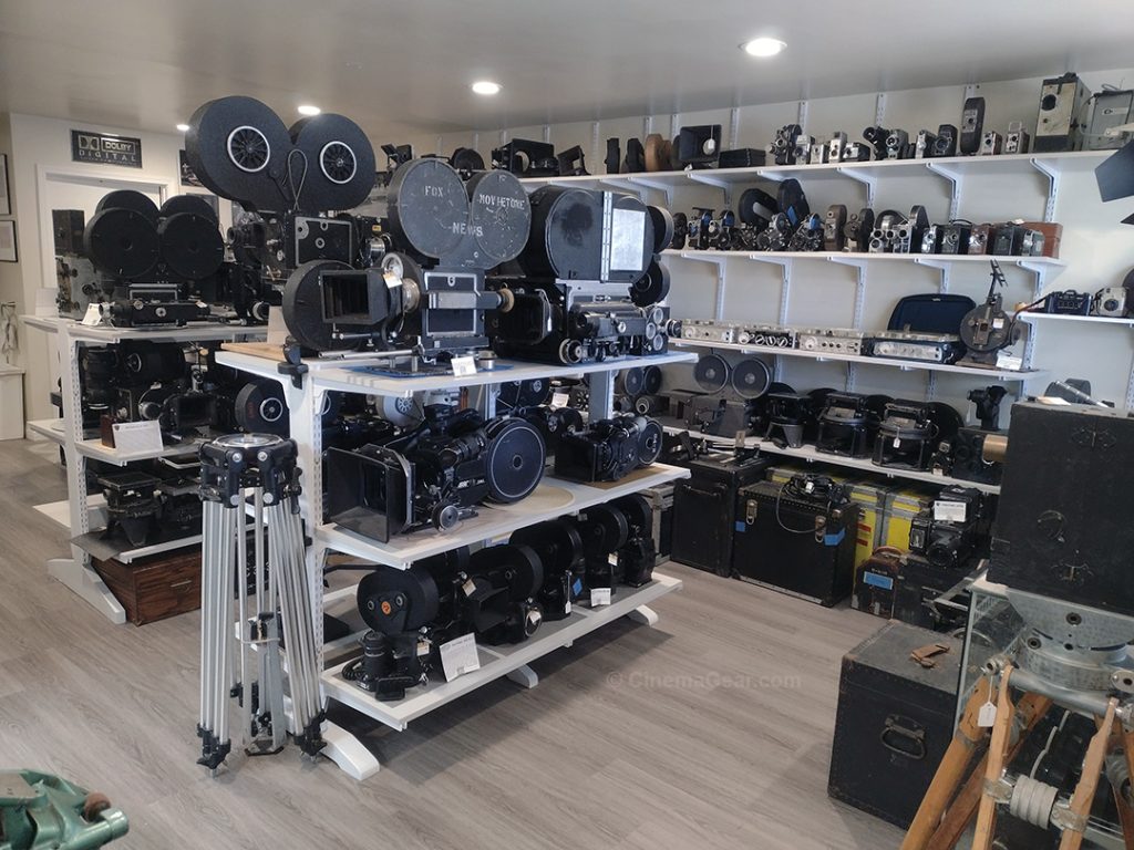 The showroom at CinemaGear looking from the front door into the room. You can see a set of wall shelves stocked with cameras, gyro heads, and Nagra tape recorders, along with two sets of free standing shelves filled with cameras made by the Mitchell Camera Corporation, Arriflex, the William P. Stein Co., and some Mitchell geared heads. A metal tripod stands in the foreground against the front shelving unit.
