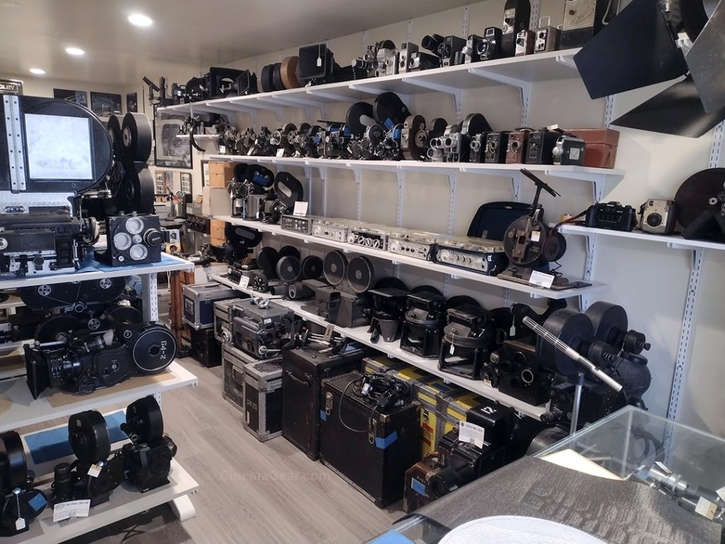 The showroom at CinemaGear looking from the front window, showing a closer look at the wall shelves. The top shelf contains a row of 8mm and 16mm cameras, a matte box, and some camera magazines. The second shelf contains a row of Bell & Howell Eyemo, Bell & Howell Filmo, Bolex, Arriflex, Victor Animatograph Co., and Kodak 16mm and 35mm motion picture cameras. The third shelf contains some Arriflex 35mm motion picture cameras, four Nagra tape recorders, one Tandberg tape recorder, and an Arriflex 16mm contact printer. The bottom shelf contains an Arriflex 35mm camera, a Mitchell 16mm SS camera and sound recording mixing console, a Maurer model 05 16mm camera, an Auricon CM-74 16mm camera, a Mitchell friction head, two Akeley Gyro heads on high hats, and an Acme Model 6 35mm camera. Below are a series of equipment cases, a partially assembled Mitchell BNCR camera, a Peter Lisand body brace, some cables, and a Mitchell KF8 aerial reconnaissance camera. You can also see the corner of our new shelving unit in the right corner, and part of a glass case that contains lenses in the foreground.